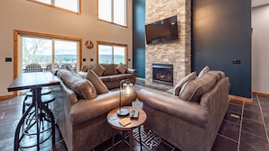 A modern living room with a stone fireplace, leather sofas, a dining table, and large windows offering a view of the outdoors.