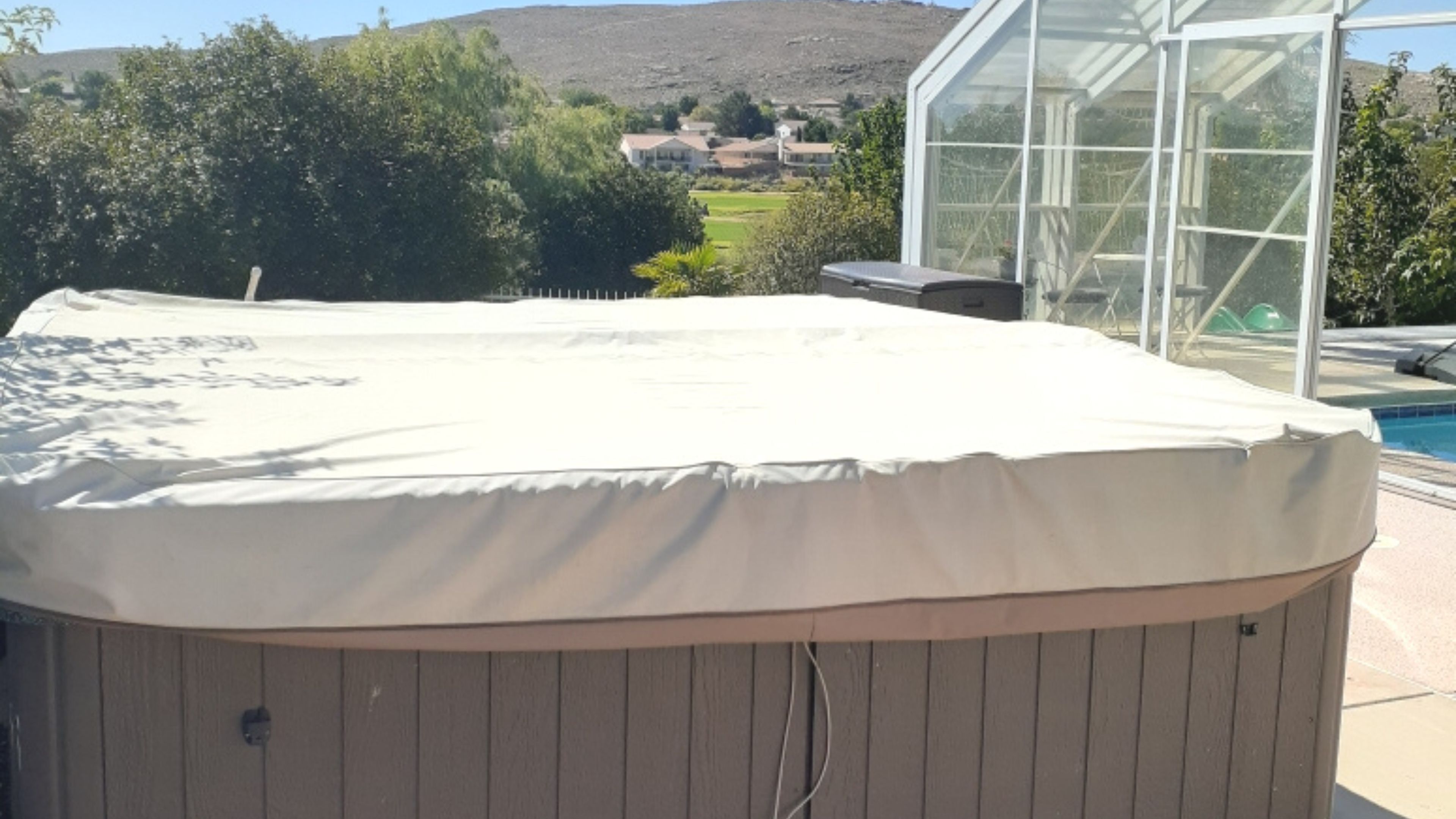 A hot tub with a cover, surrounded by a wooden deck, a pool, and a greenhouse structure.