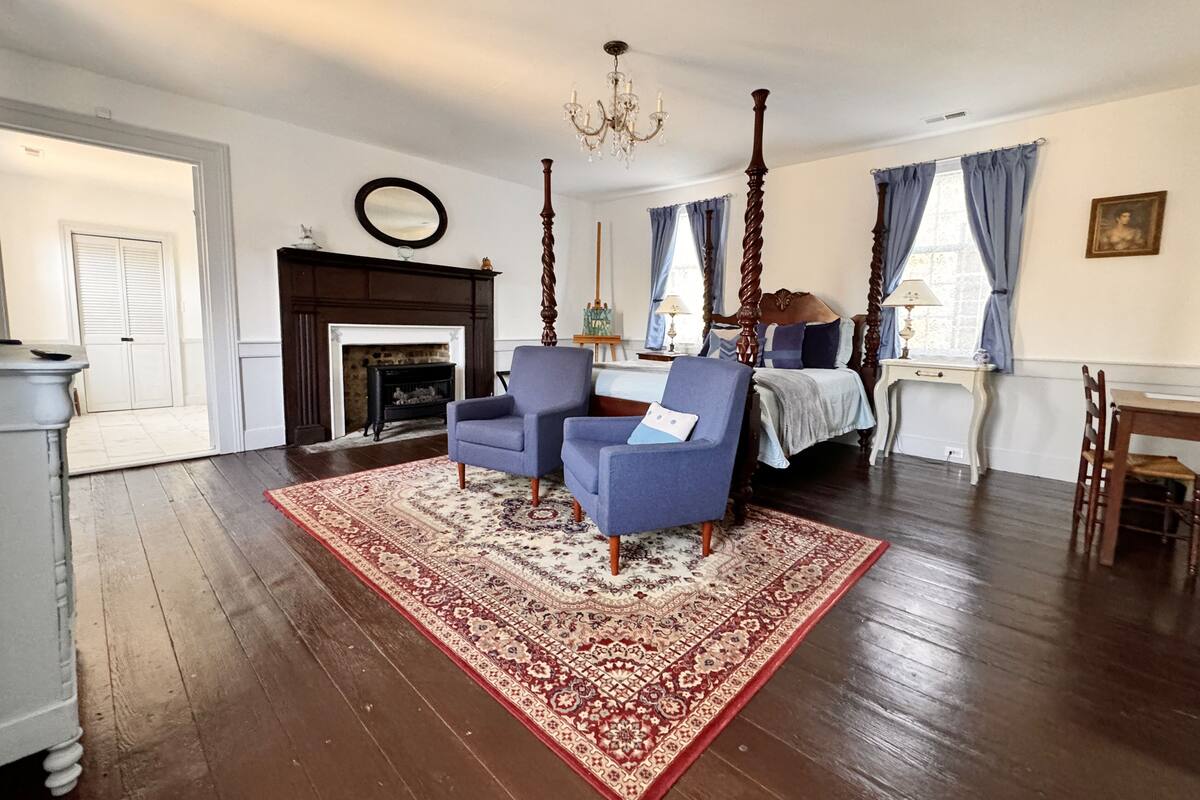 Una habitación amplia con piso de madera, cama con dosel, dos sillones, chimenea y candelabro.