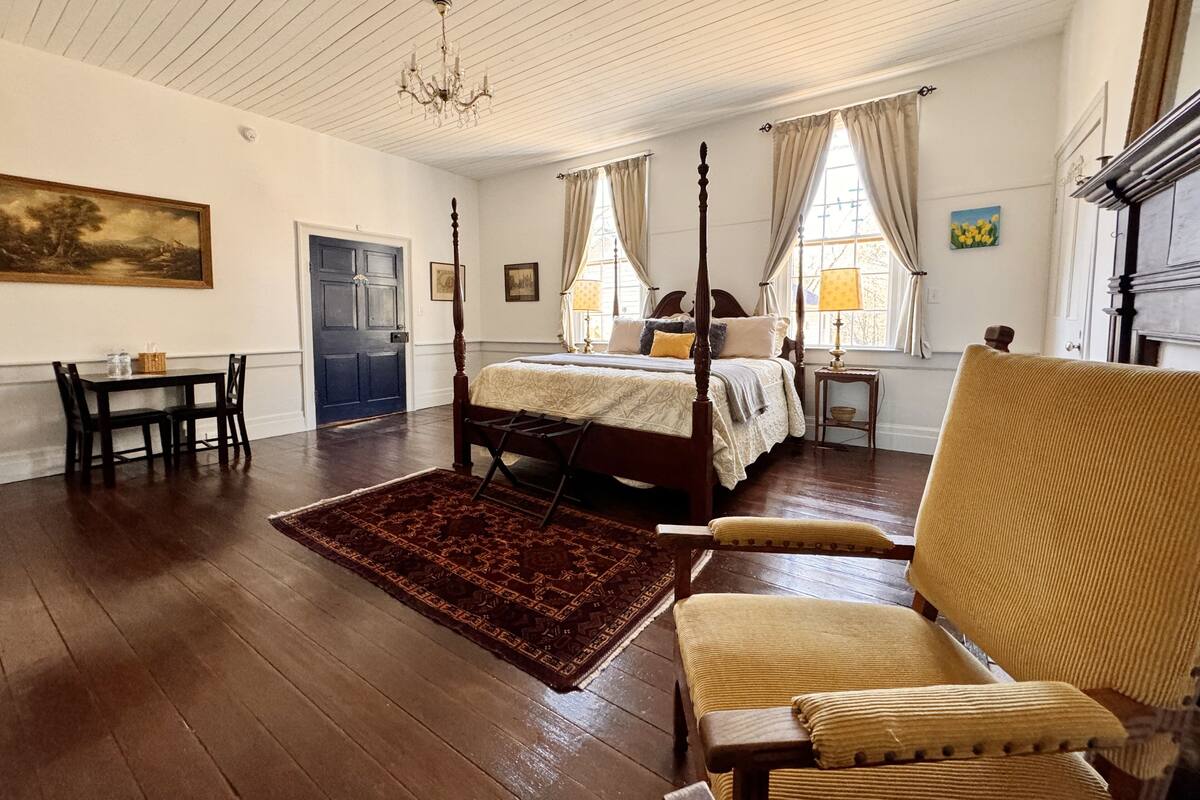 Un dormitorio espacioso con cama con dosel, una silla de madera, una mesita y un cuadro en la pared.