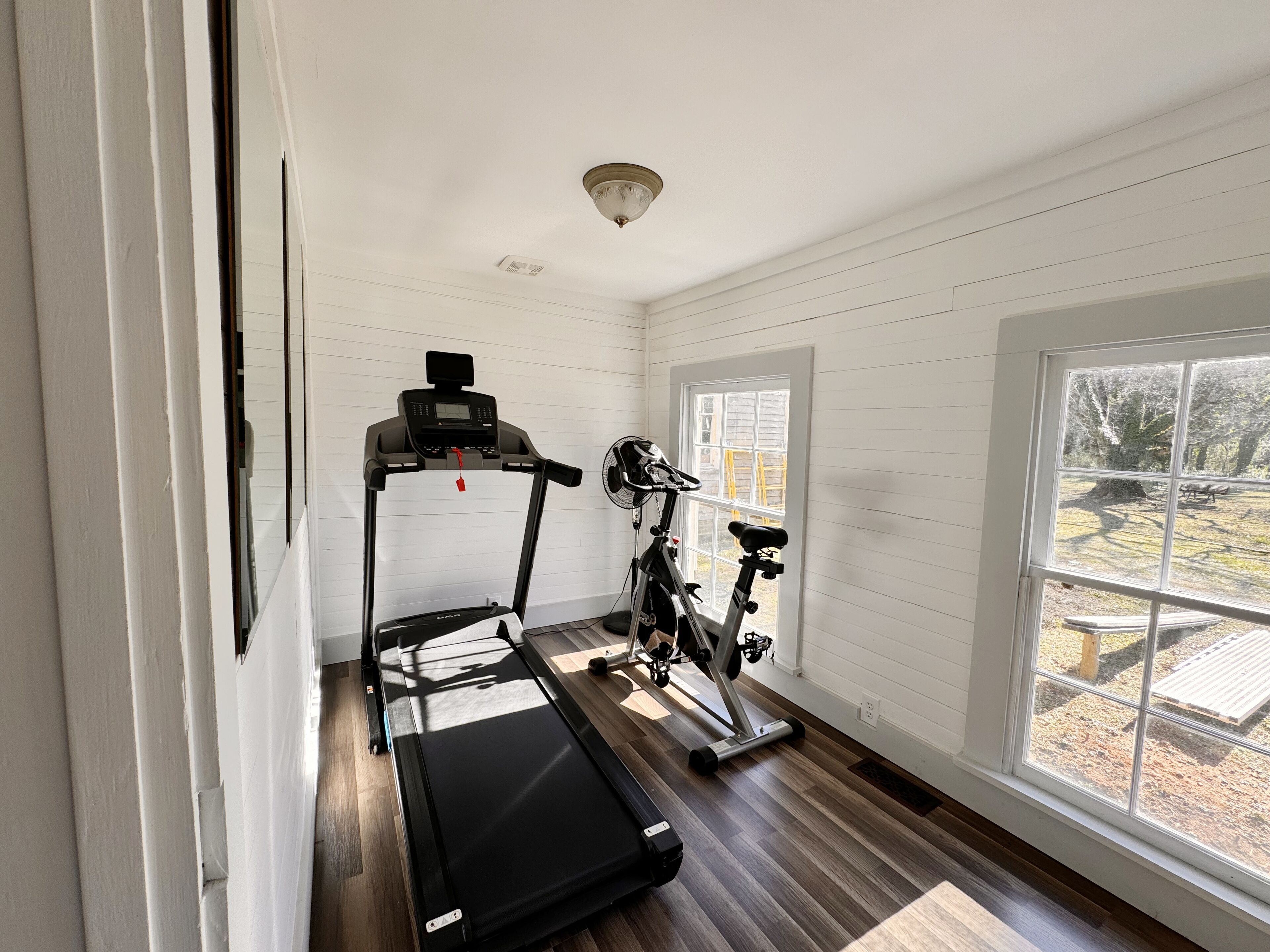 A room with a treadmill, stationary bike, and a window with a view of a park.