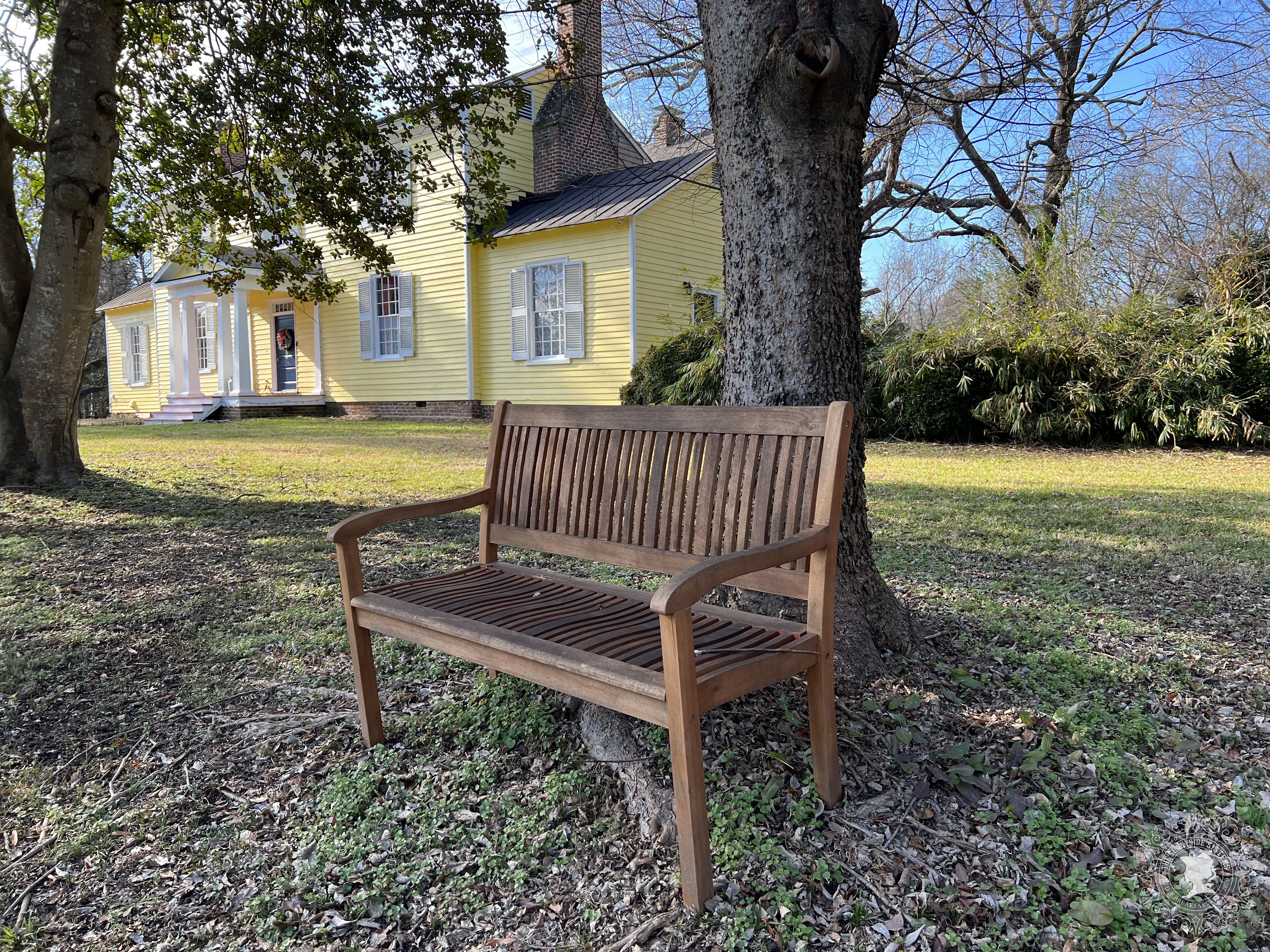 Un banco de madera en un parque, con una casa amarilla al fondo.