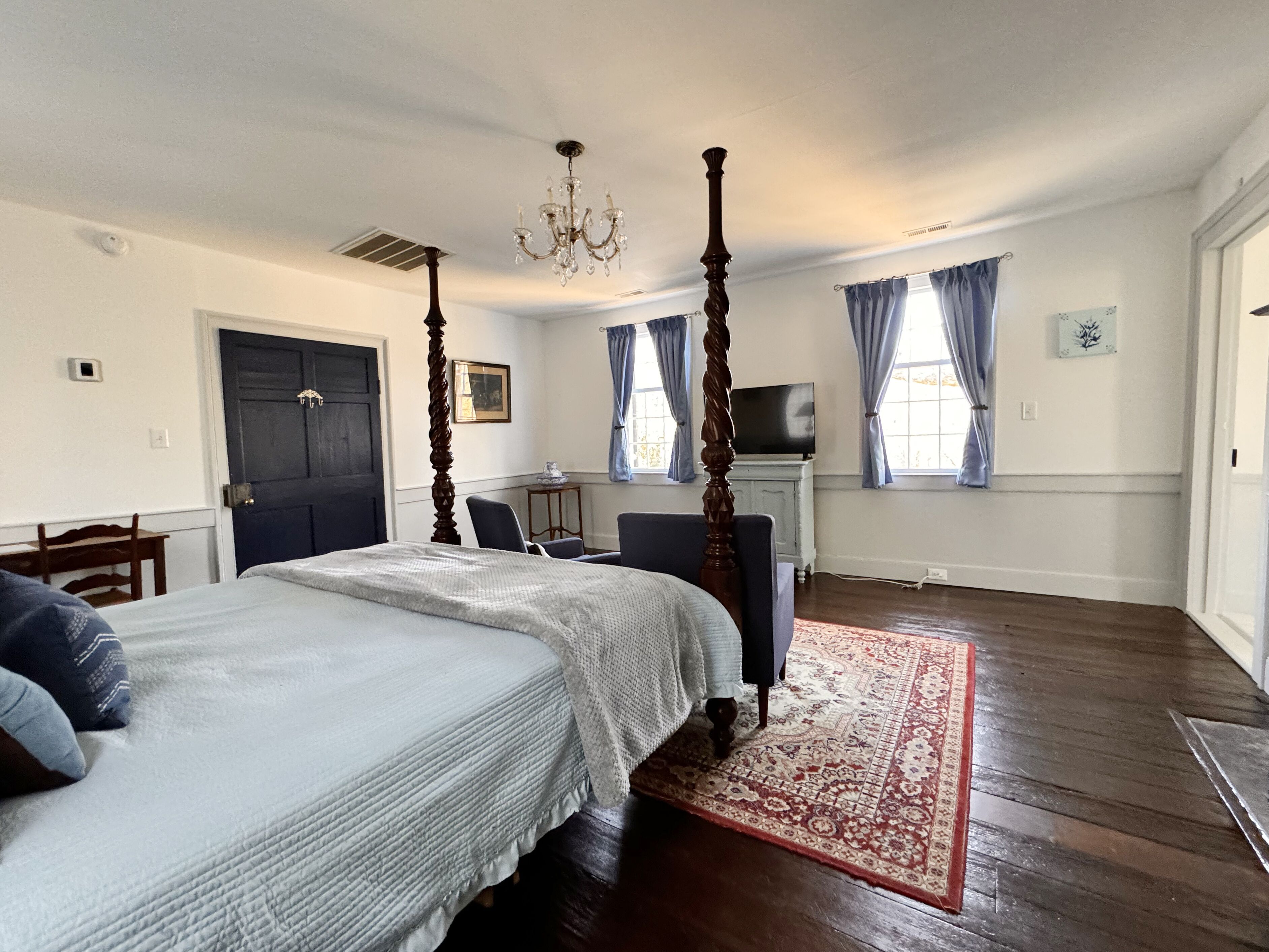 A bedroom with a four-poster bed, a chandelier, a television, and a large window with curtains.