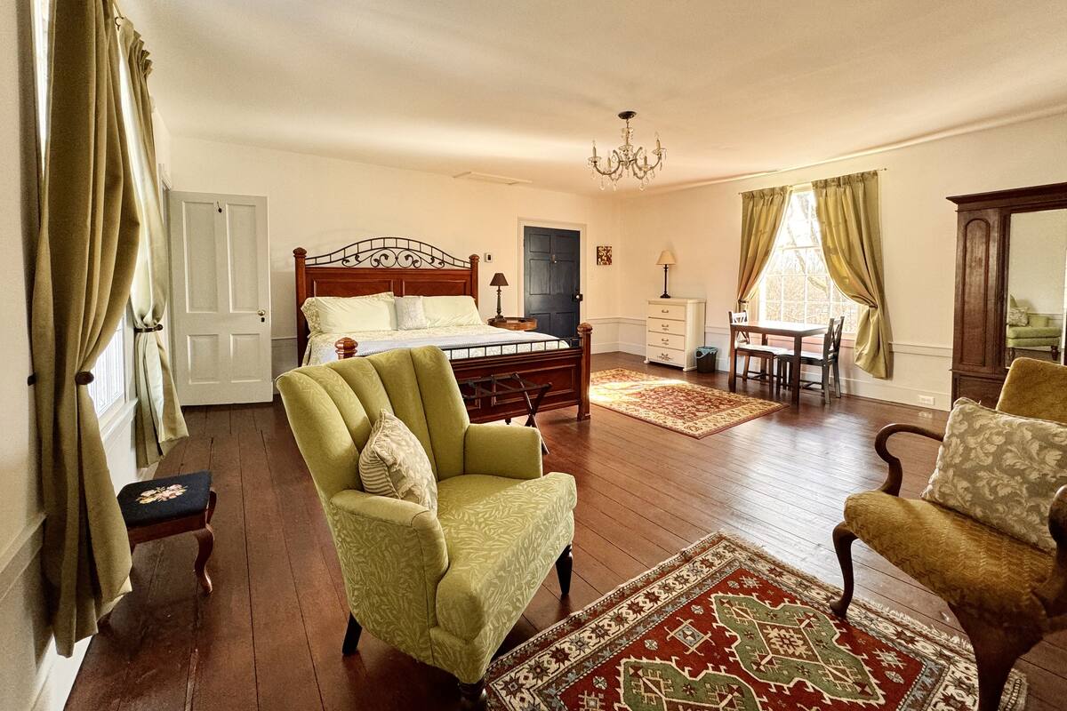Un dormitorio con cama, dos sillones, cómoda, mesita de noche y ventana con cortinas.