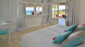 A bedroom with a large bed, a small table, and a chair. There is a view of the ocean through the open window.