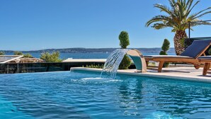 A pool with a waterfall feature, surrounded by lounge chairs and palm trees, overlooking a coastal landscape. - Luxury Villa Imperio with Pool (Kaštel Gomilica)