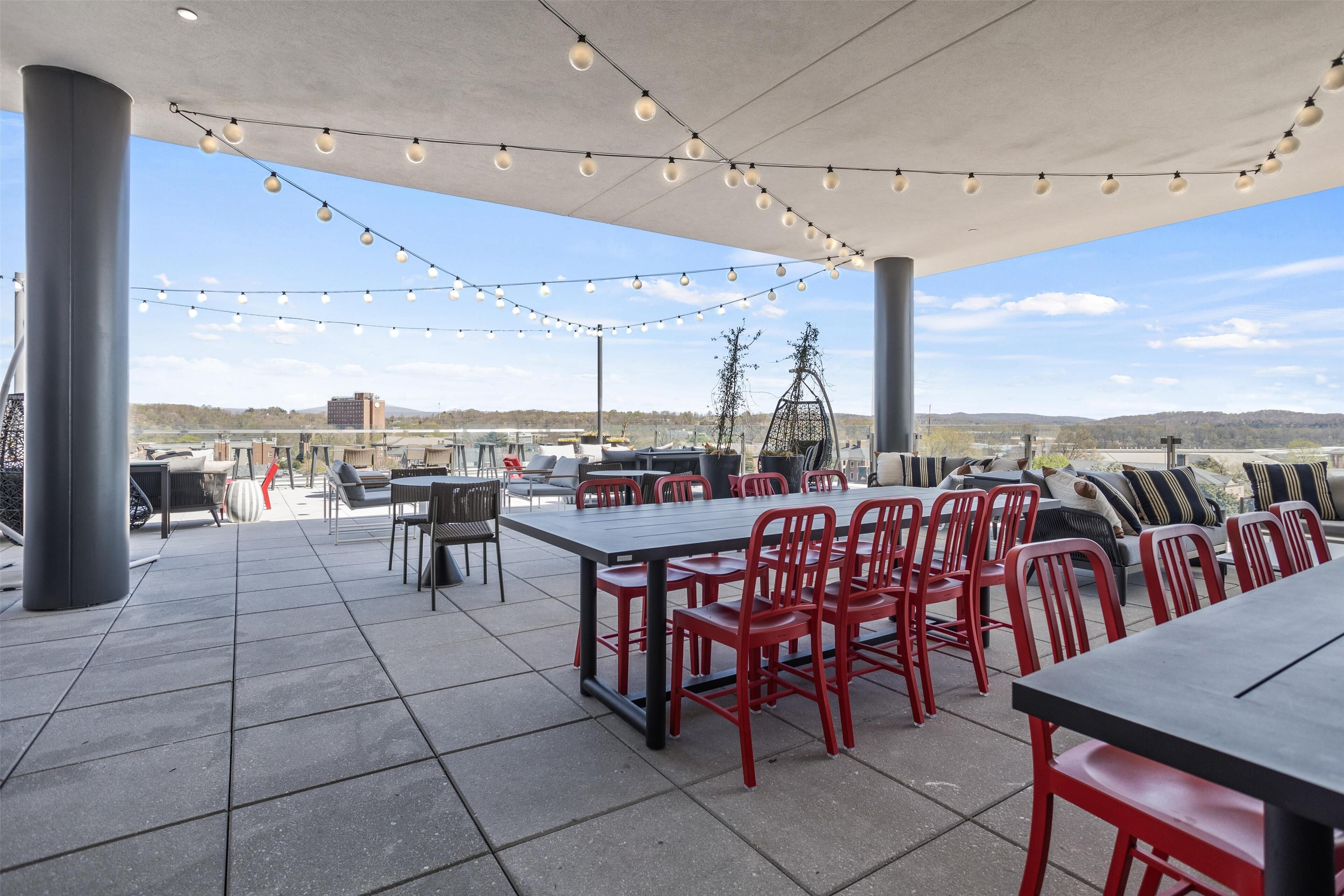An outdoor patio with a long table and red chairs, string lights overhead, and a view of distant hills.