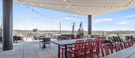 An outdoor patio with a long table and red chairs, string lights overhead, and a view of distant hills.