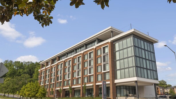 A modern multi-story building with large windows and a flat roof, surrounded by greenery and a clear sky.