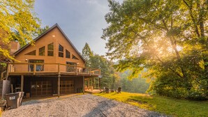 Une maison en bois à deux étages, dotée d’un balcon, d’une allée en gravier et entourée d’une zone boisée.