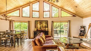 Un salon spacieux avec un plafond haut, de grandes fenêtres, un canapé en cuir, une table basse en verre et un coin repas avec une table et des chaises en bois.
