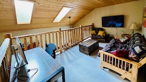 Um quarto estilo loft acolhedor, com teto de madeira, teto inclinado com claraboias, sofá de madeira, uma mesa pequena e uma TV.