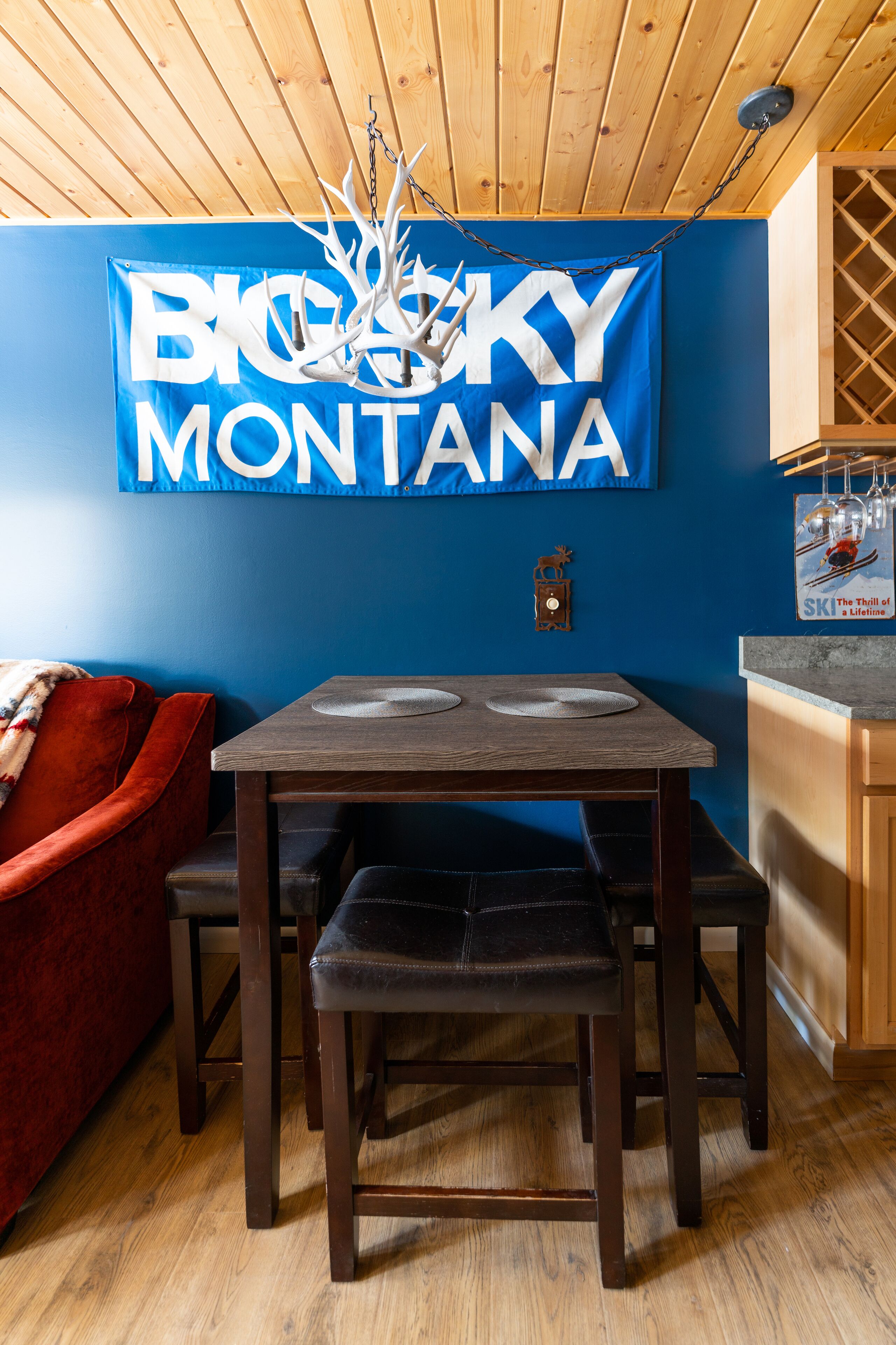A dining area with a wooden table and chairs, a blue wall with a 'Big Sky Montana' banner, and a red couch.