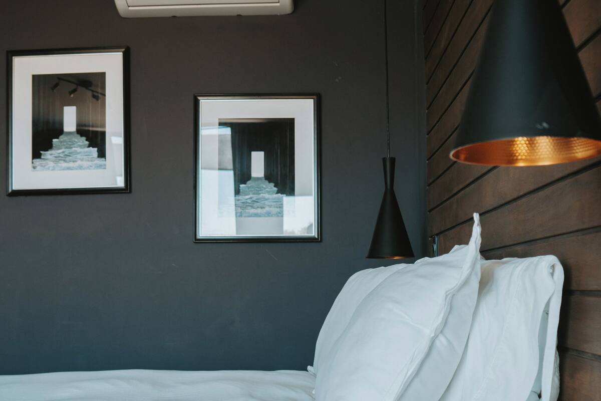 Ein Schlafzimmer mit einem Bett, zwei gerahmten Bildern an der Wand, einer schwarzen Pendelleuchte und einer Wand-Klimaanlage.