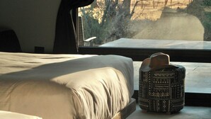 A bed with a white sheet and a patterned ottoman in front of a window with a view of trees and a dirt path. - Pulso Valle de Guadalupe (Valle de Guadalupe)