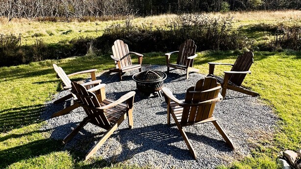 Un espace extérieur aménagé avec des chaises Adirondack et un foyer entouré de gravier, le tout se fondant dans un décor composé d’arbres et d’un ciel dégagé.