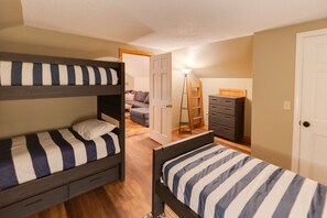 A dorm-style room with bunk beds, a dresser, a lamp, and a door leading to another room. - Loon Mountain Condo- Walk to Main Street! (Lincoln)