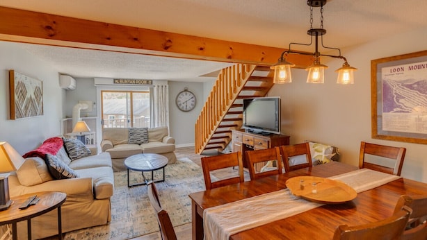 A cozy living room with a sofa, coffee table, and dining table. There's a TV, a clock, and a staircase leading to another level. - Loon Mountain Condo- Walk to Main Street! (Lincoln)