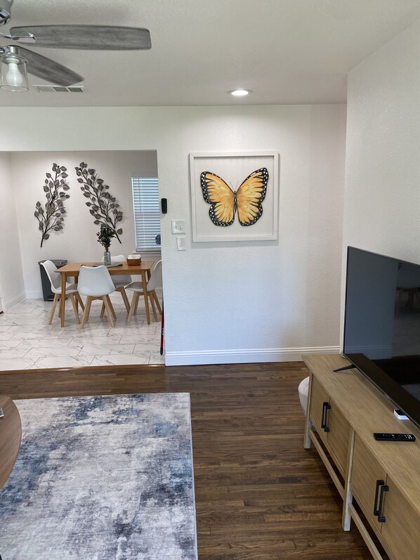 Een moderne woonkamer met een houten tv-meubel, een groot schilderij van een vlinder en een eethoek met houten stoelen en een tafel.