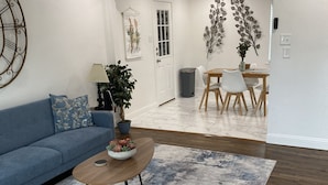 A modern living room with a blue sofa, a wooden coffee table with a plant, a patterned rug, and a dining area with a wooden table and chairs.