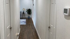 A hallway with white walls and wooden flooring, leading to a room with a lamp and a potted plant.