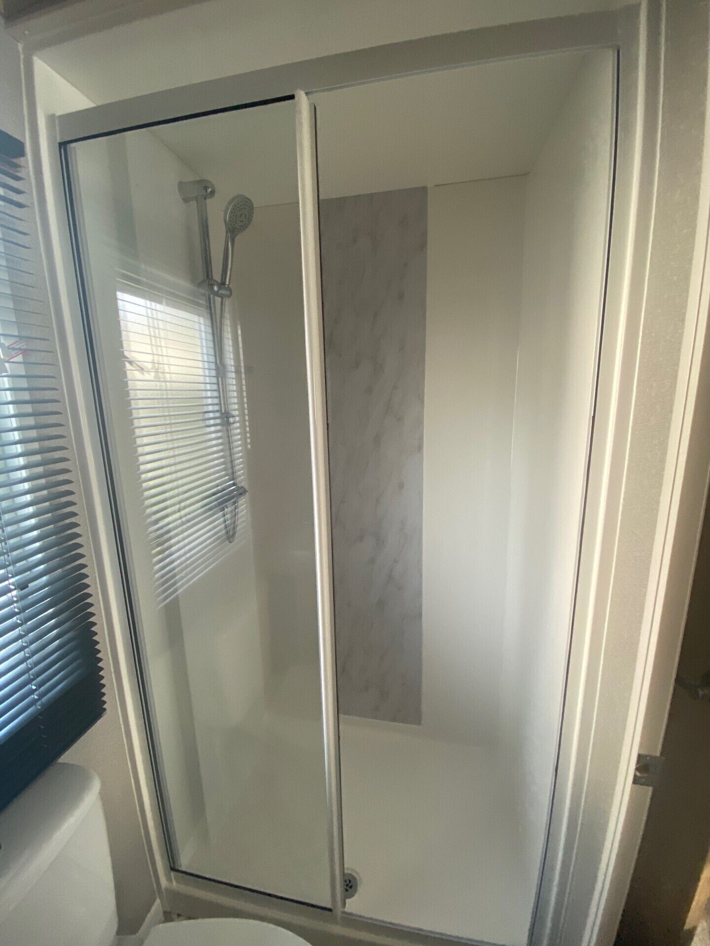 A glass-enclosed shower with a marble wall and a window with blinds.