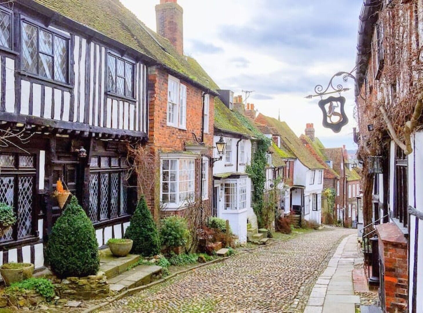 A narrow cobblestone street lined with traditional timber-framed houses, each with distinct architectural features and small front gardens.