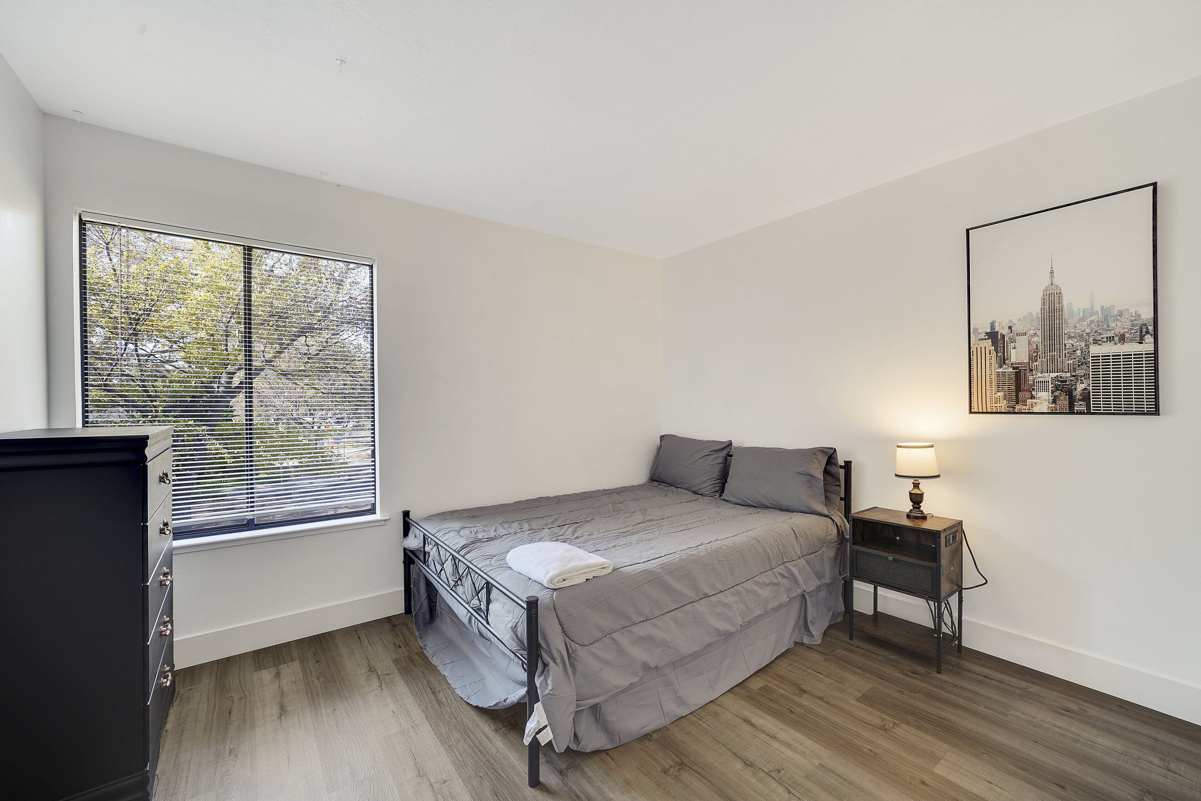 A bedroom with a bed, a nightstand, a window with blinds, a dresser, and a cityscape painting on the wall.