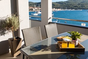 Ein Balkon mit Glastisch, Korbstühlen, einem Tablett mit Gläsern und einer Pflanze, sowie Blick auf das Meer und Boote.