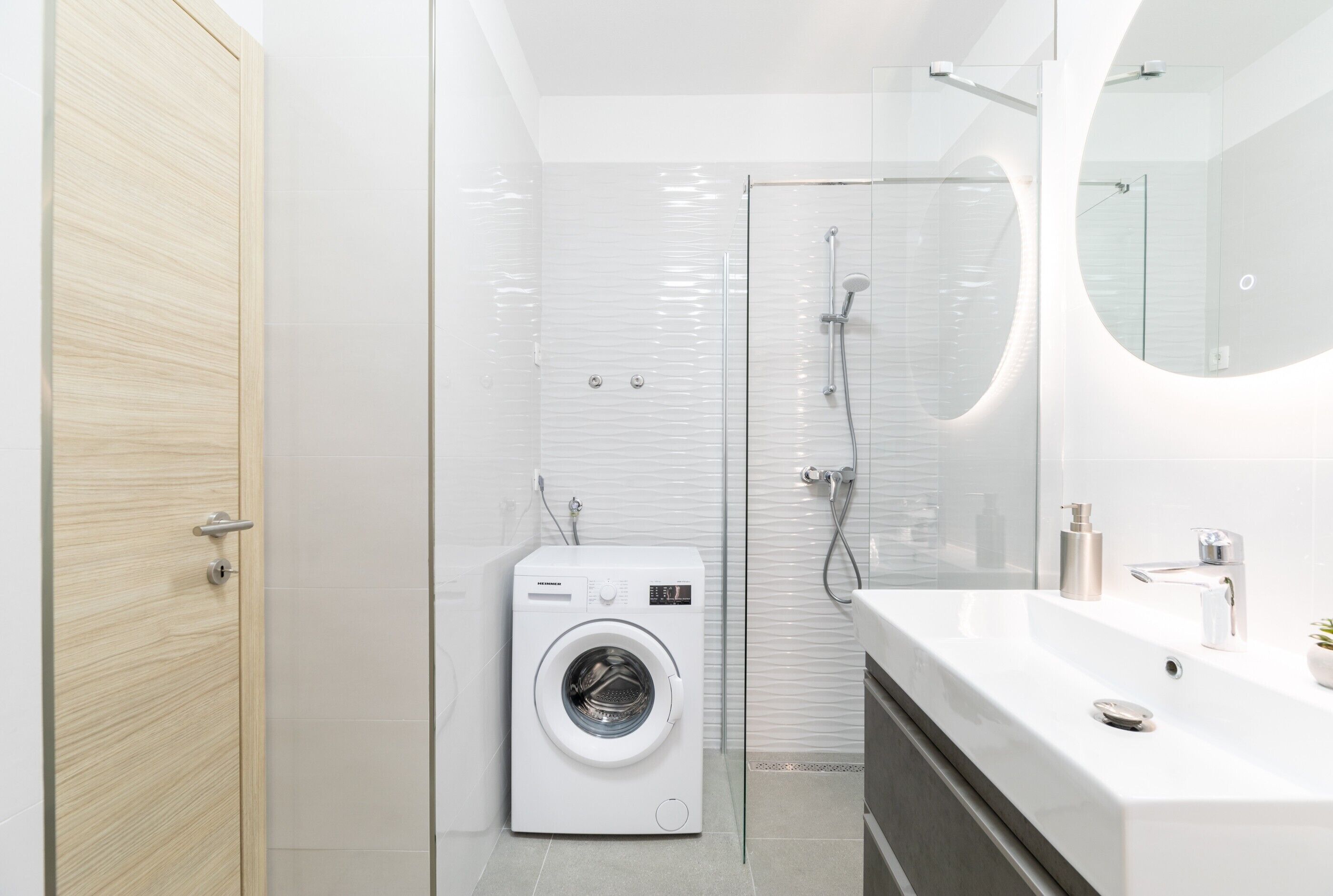 A modern bathroom with a built-in washing machine, a glass shower enclosure, and a large mirror above the sink.