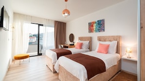 Ein Hotelzimmer mit zwei Betten, einem Sofa, einem kleinen Tisch, einem Fernseher, einem Gemälde und Blick auf die Stadt.