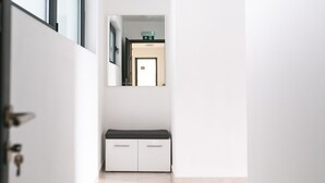 Un pasillo con un banco, una puerta y un espejo que refleja una habitación con cama y ventana.