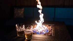 Ein Tisch mit einem kleinen Feuer, zwei Biergläsern und einem gemusterten Kissen im Hintergrund.