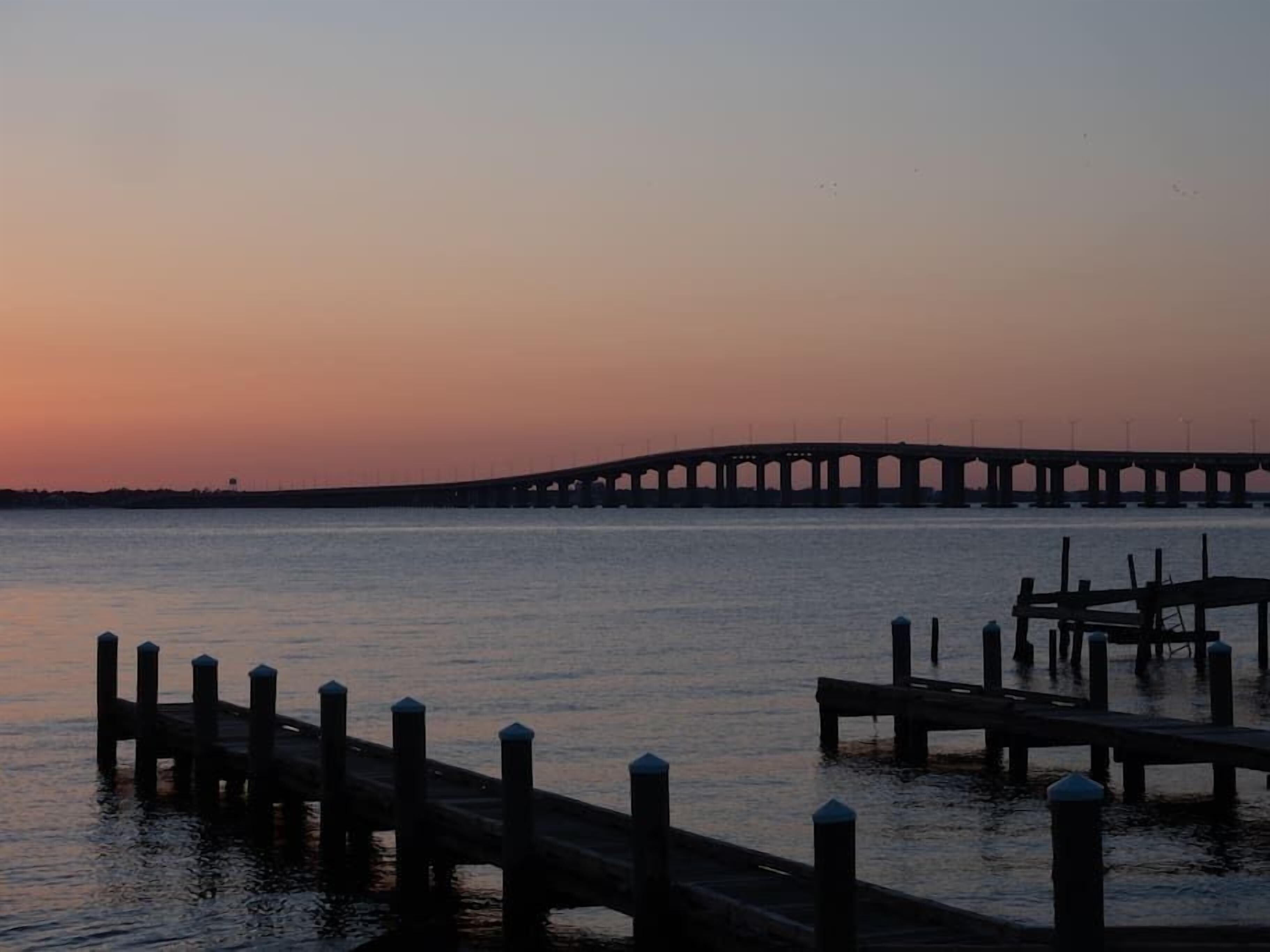 A pier extending into calm water with a bridge in the background.