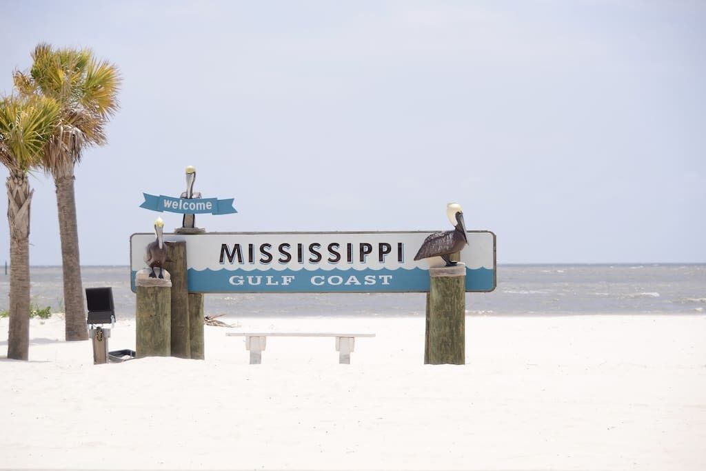 Un cartel de bienvenida a la Costa del Golfo de Mississippi, con un banco y palmeras al fondo.