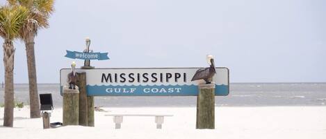 Un cartel de bienvenida a la Costa del Golfo de Mississippi, con un banco y palmeras al fondo.