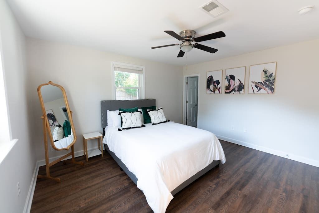 A bedroom with a bed, a ceiling fan, a mirror, a window, and wall art.