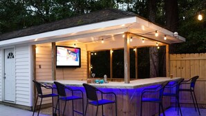 Un área de bar al aire libre con estructura de madera, taburetes de bar y una pantalla de televisión.