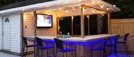 An outdoor bar area with a wooden structure, bar stools, and a television screen.