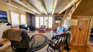 Una sala de estar acogedora con chimenea, vigas de madera, un sofá, un sillón mecededor y una escalera.