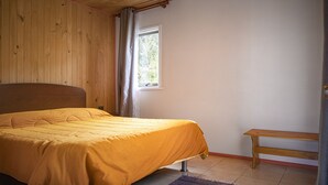 A bedroom with a wooden headboard, a single bed with a yellow bedspread, a window with curtains, a wooden bench, and a tiled floor.