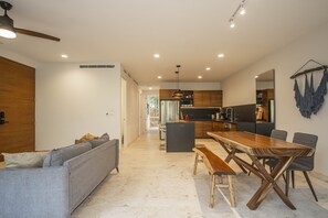 A modern kitchen with a dining area, featuring a wooden dining table and benches, a grey sofa, and a ceiling fan.