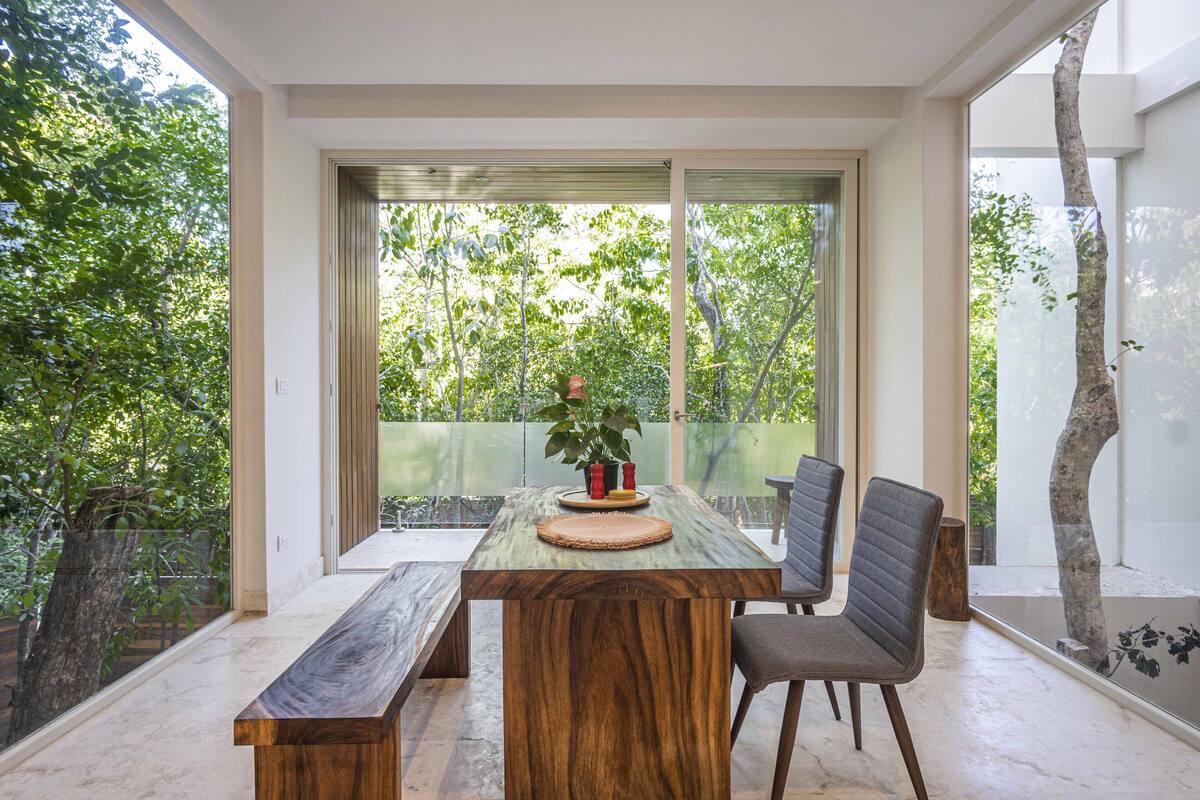 Un comedor moderno con una mesa de madera, dos sillas y un banco. Amplias ventanas ofrecen vistas al verde exterior.