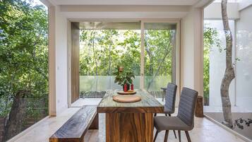 Un comedor moderno con una mesa de madera, dos sillas y un banco. Amplias ventanas ofrecen vistas al verde exterior.