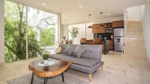 A modern living room with a grey sofa, wooden coffee table, and large windows offering a view of trees. - Vita Tulum (Tulum)