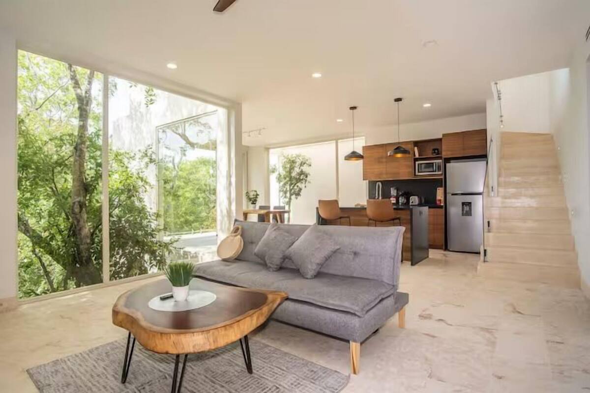 Una sala de estar moderna con un sofá gris, una mesa de centro de madera y grandes ventanas que dan a una vista de árboles.