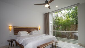 A bedroom with a large bed, a wooden headboard, bedside lamps, a bench, and a view of greenery through a large window. - Vita Tulum (Tulum)