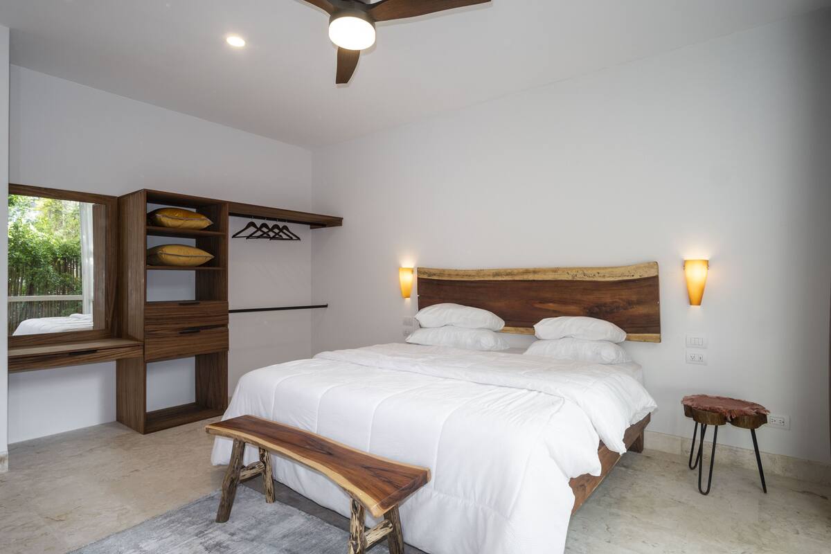 Un dormitorio con una cama grande, un banco de madera y un estante empotrado con ropa colgada.