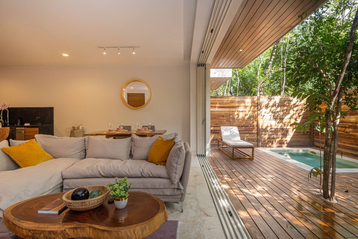 Una sala moderna con un sofá, una mesa de centro y un televisor. Hay una terraza de madera con un jacuzzi y un árbol.