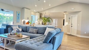 A modern living room with a sectional sofa, a coffee table, and a kitchen in the background. - SunCoast Luxury Estates-New, Spacious,  Large Heated Pool, 10 min to the Beach  (Venice)